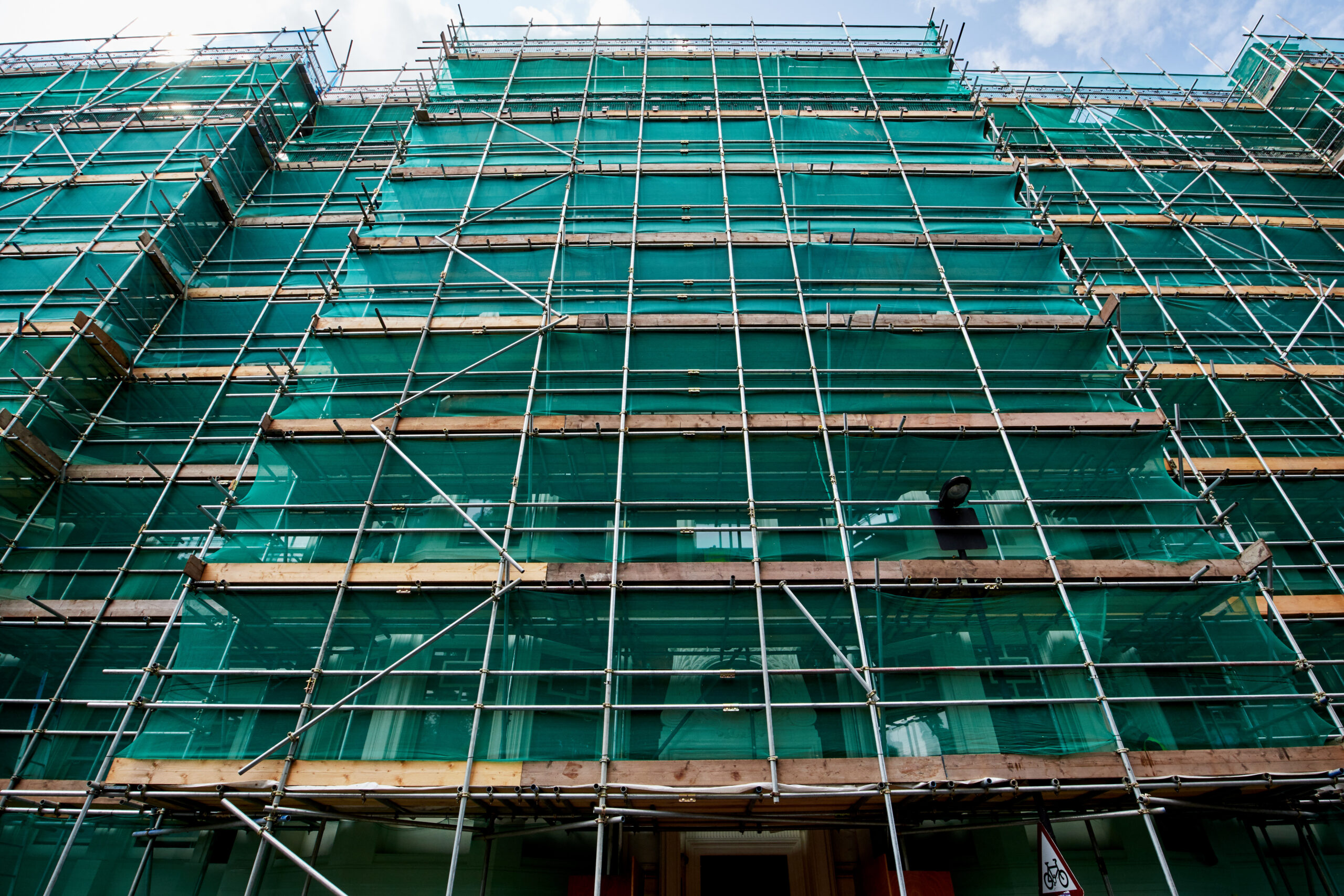 A tall building covered in green construction netting and surrounded by metal scaffolding, viewed from below, with a partly cloudy sky in the background.