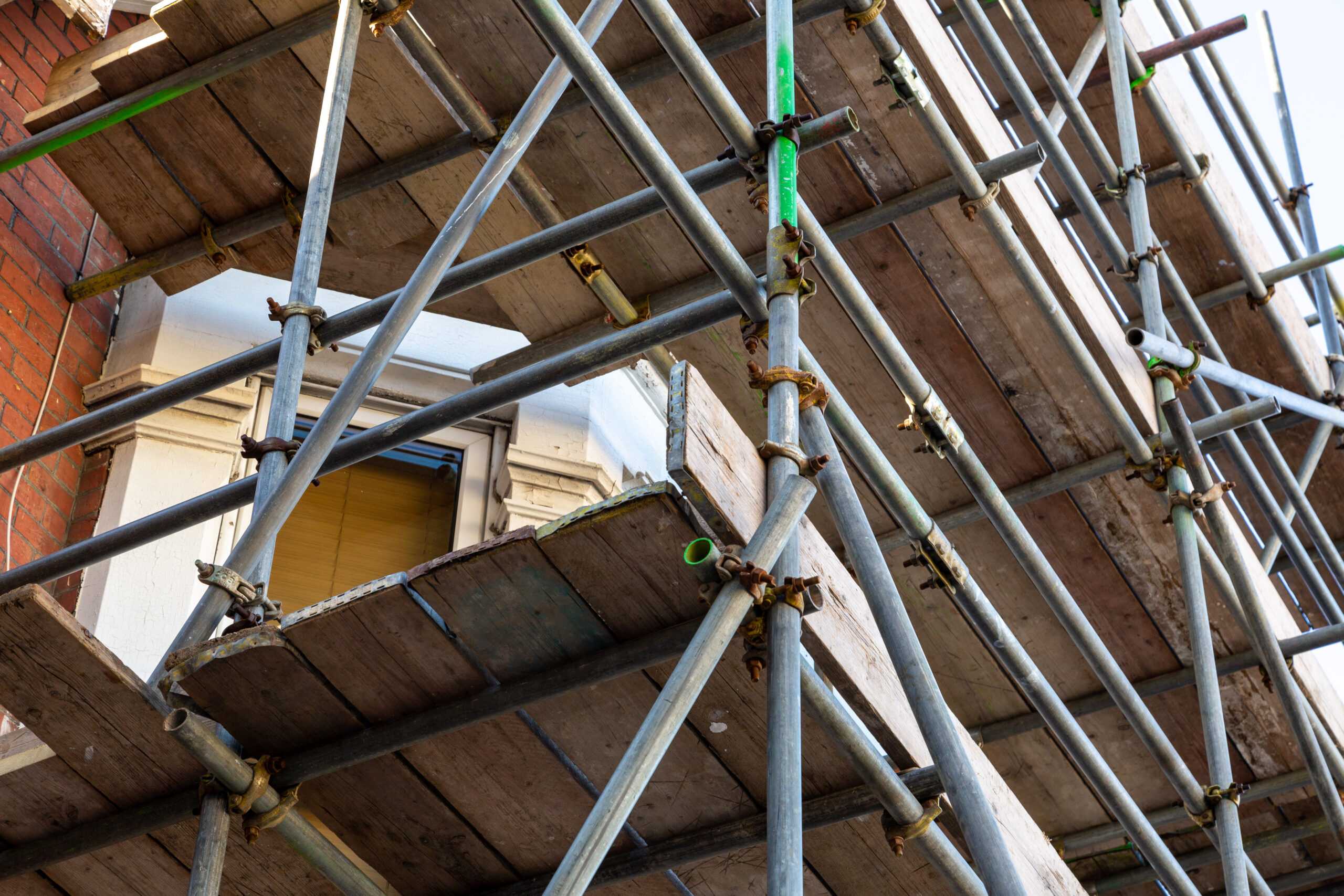 Scaffolding made of metal poles and wooden planks surrounds a building with a partially visible window and red brick wall, indicating ongoing construction or maintenance work.