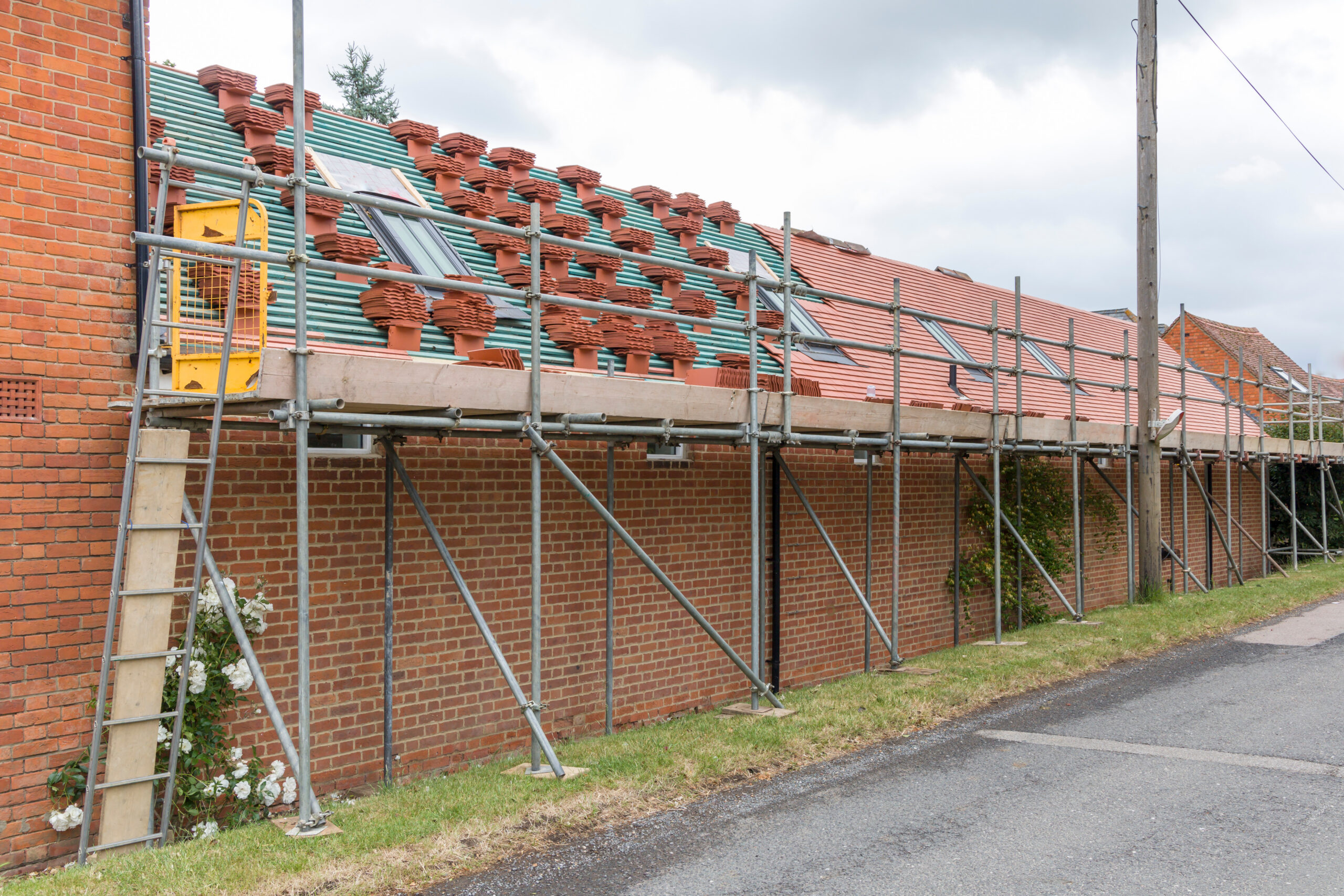 Scaffolding is set up alongside a long brick building with workers' equipment and stacks of roof tiles on a partially tiled sloped roof under a cloudy sky.