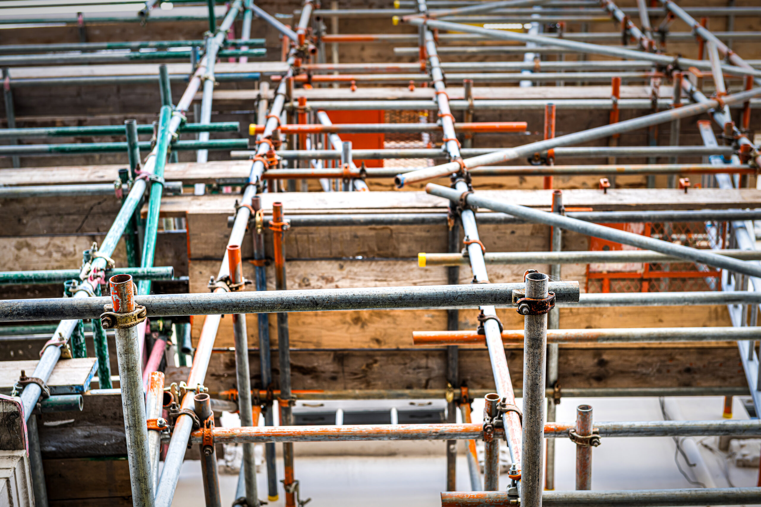 Close-up view of interconnected metal scaffolding pipes and wooden planks, showing a complex grid structure used for construction or building maintenance. The pipes and joints display signs of wear and rust.
