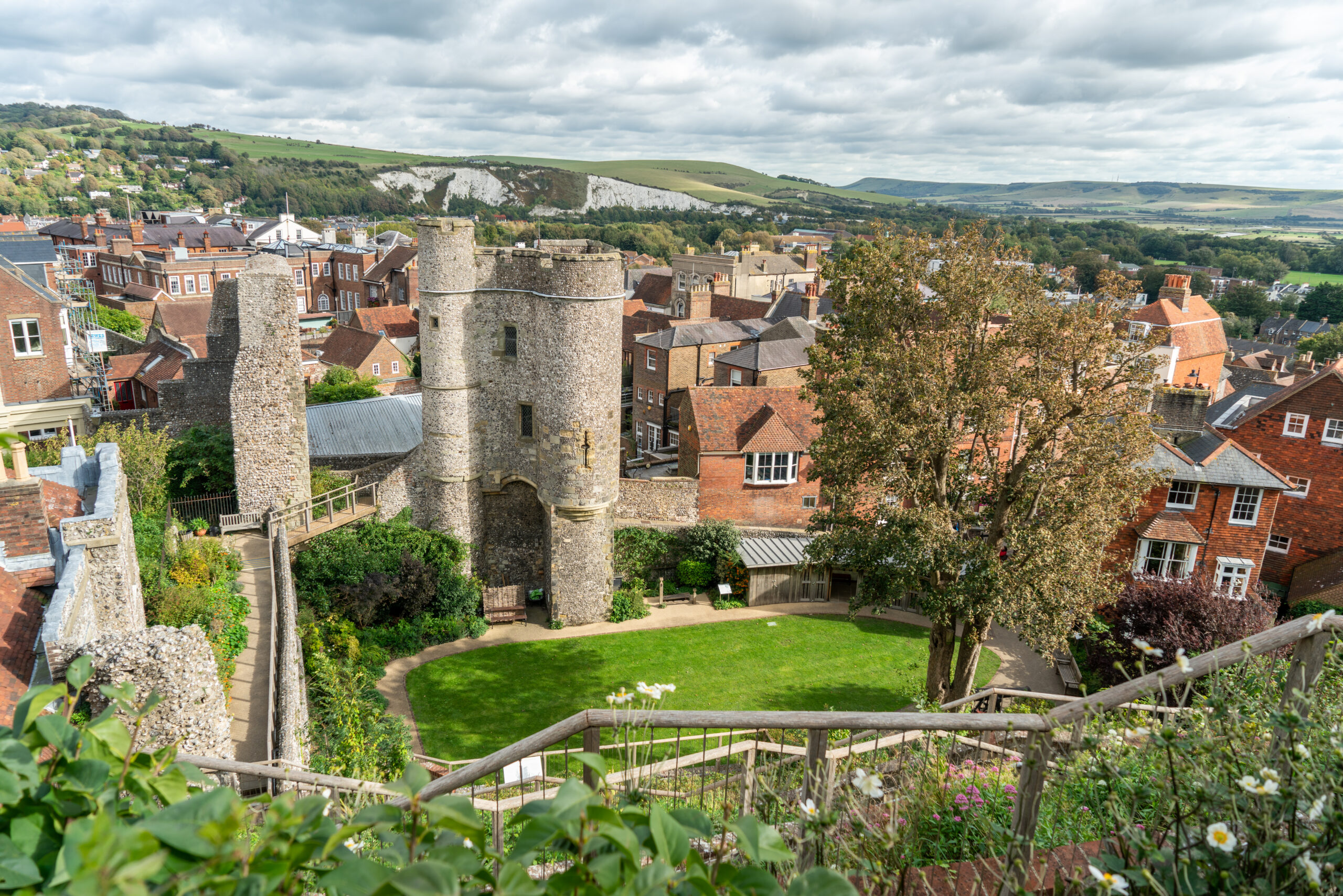 View of a medieval stone gate and towers surrounded by red-brick buildings, gardens, and green hills under a cloudy sky in a historic town.