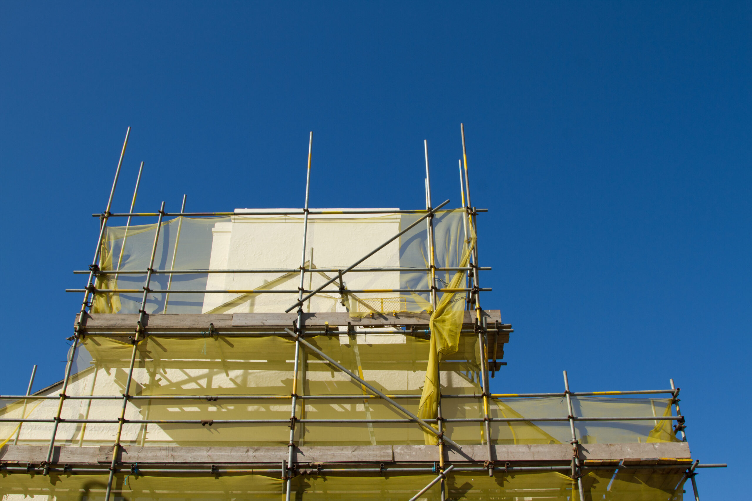 A building covered in yellow protective netting and surrounded by metal scaffolding is set against a clear blue sky.