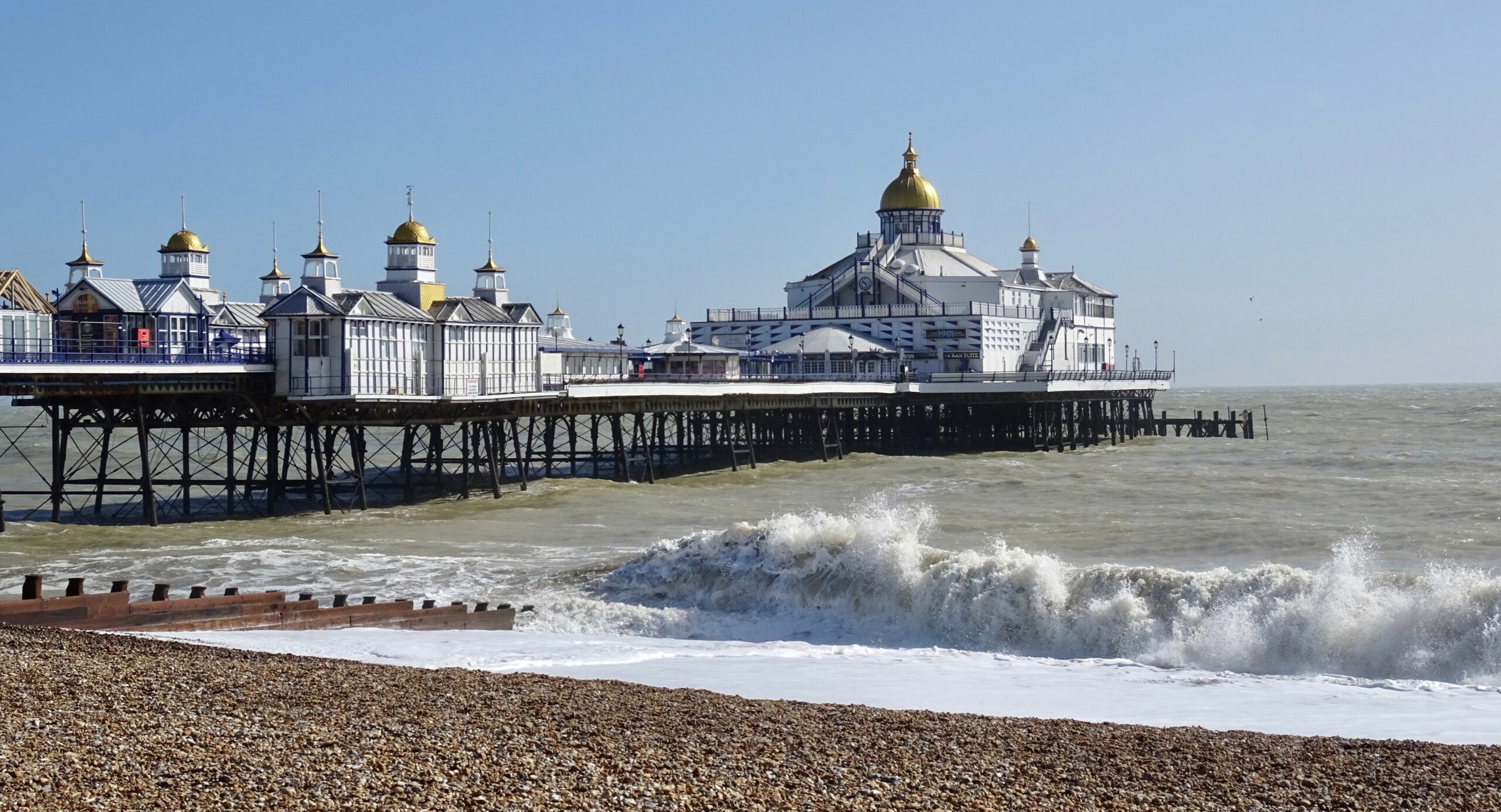 A seaside pier with white buildings and gold domes extends over the water; waves crash against the shore in the foreground under a clear blue sky.