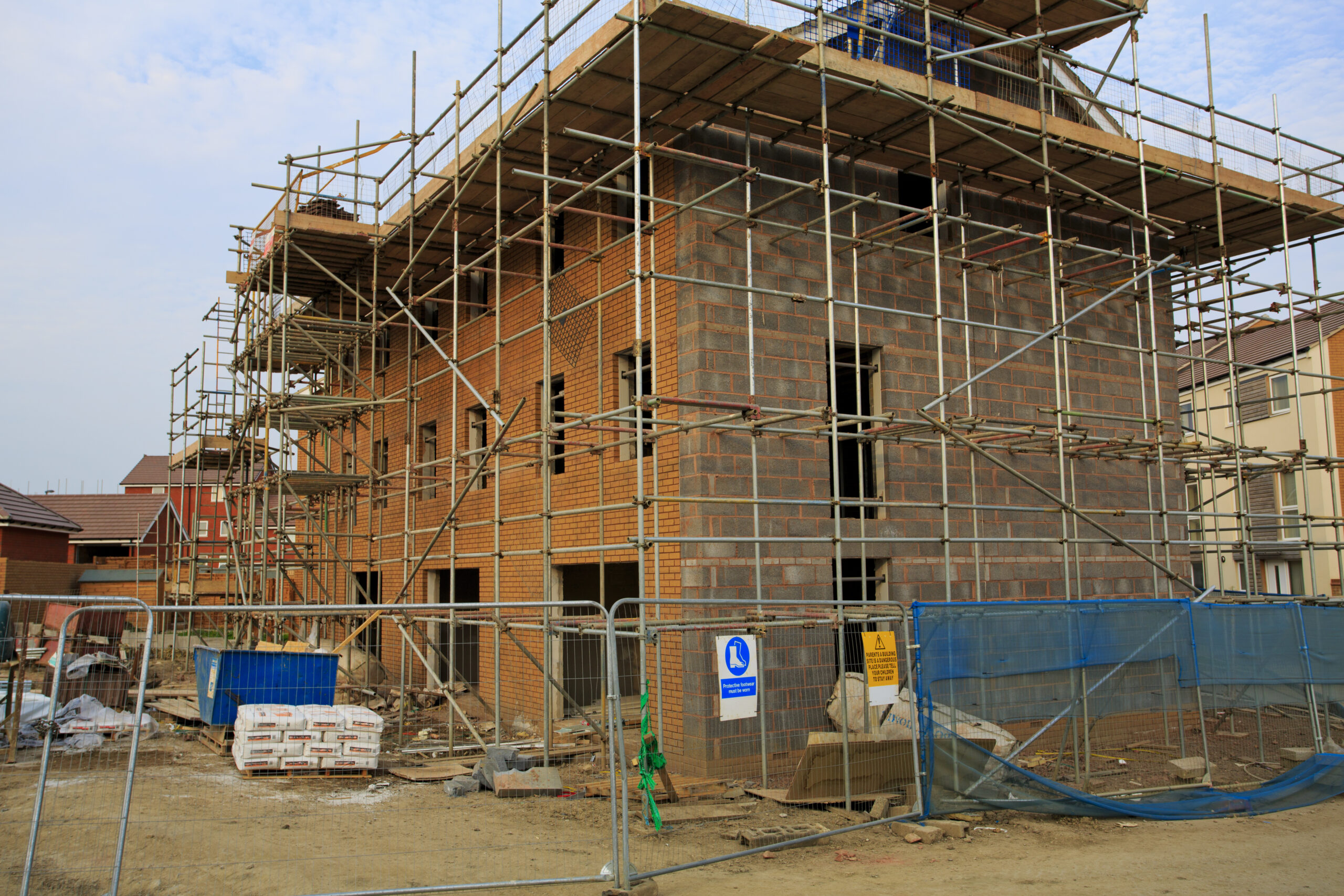 A three-story brick building under construction, surrounded by scaffolding and fencing. Construction materials, debris, and safety signs are visible on the ground. The sky is partly cloudy.