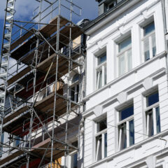 A building under renovation is partially covered with scaffolding, next to a white-painted building with large windows, under a blue sky with some clouds.