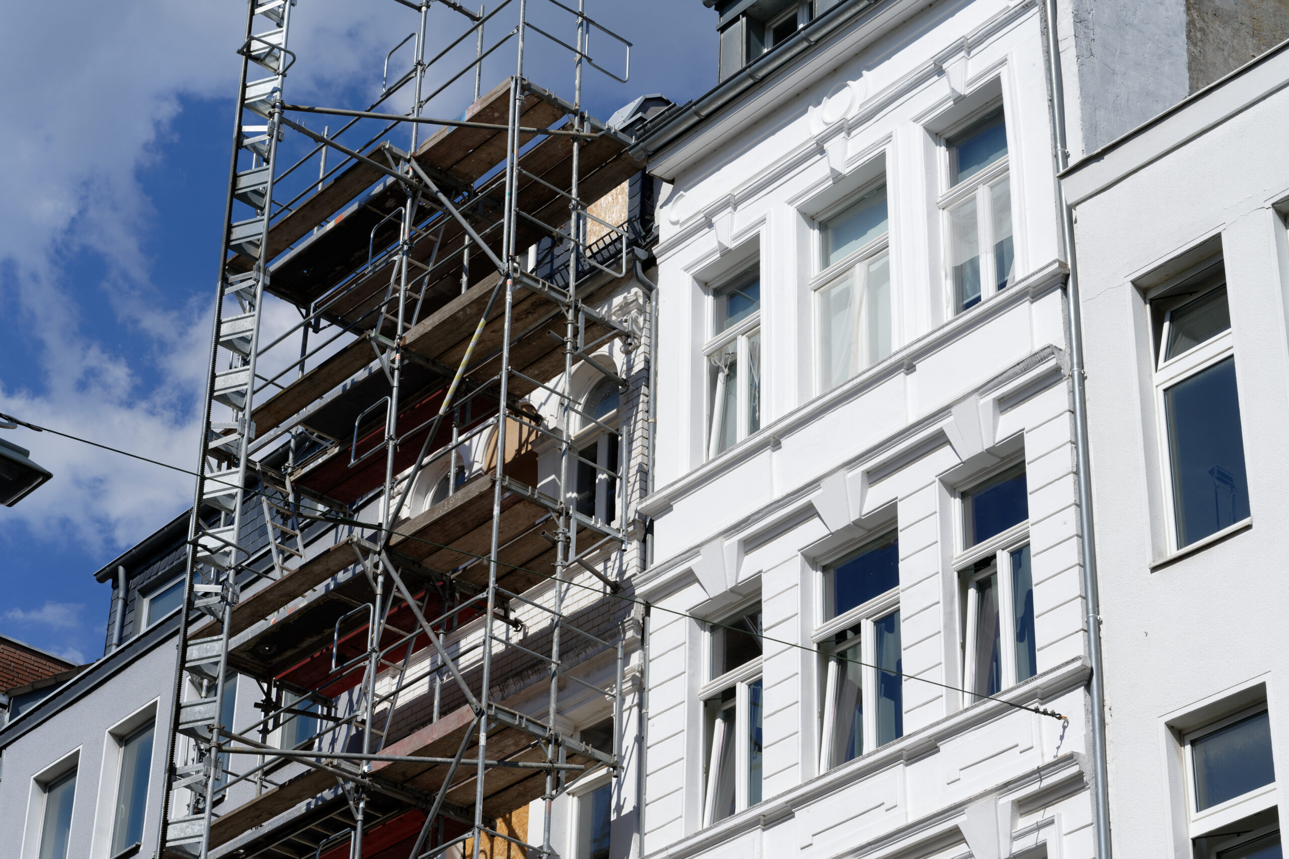 A building under renovation is partially covered with scaffolding, next to a white-painted building with large windows, under a blue sky with some clouds.