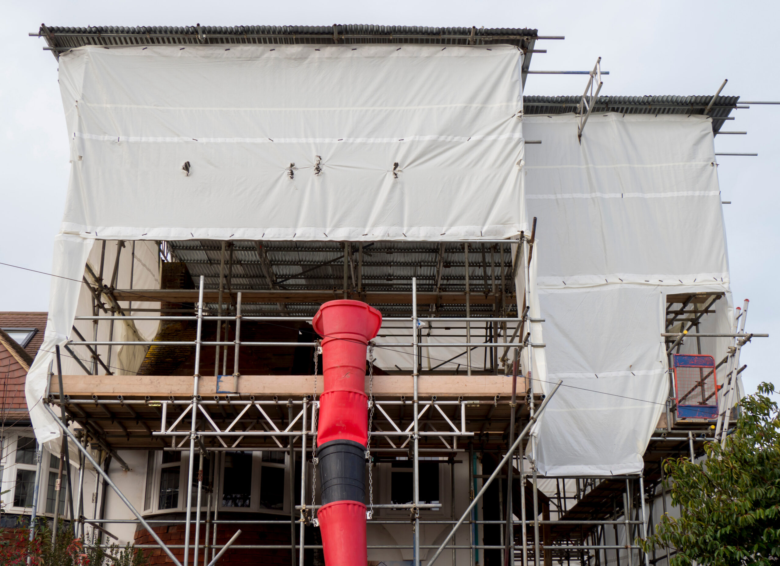 A house under construction, covered with white sheeting and surrounded by scaffolding. A red waste chute extends from the upper level down to the ground.