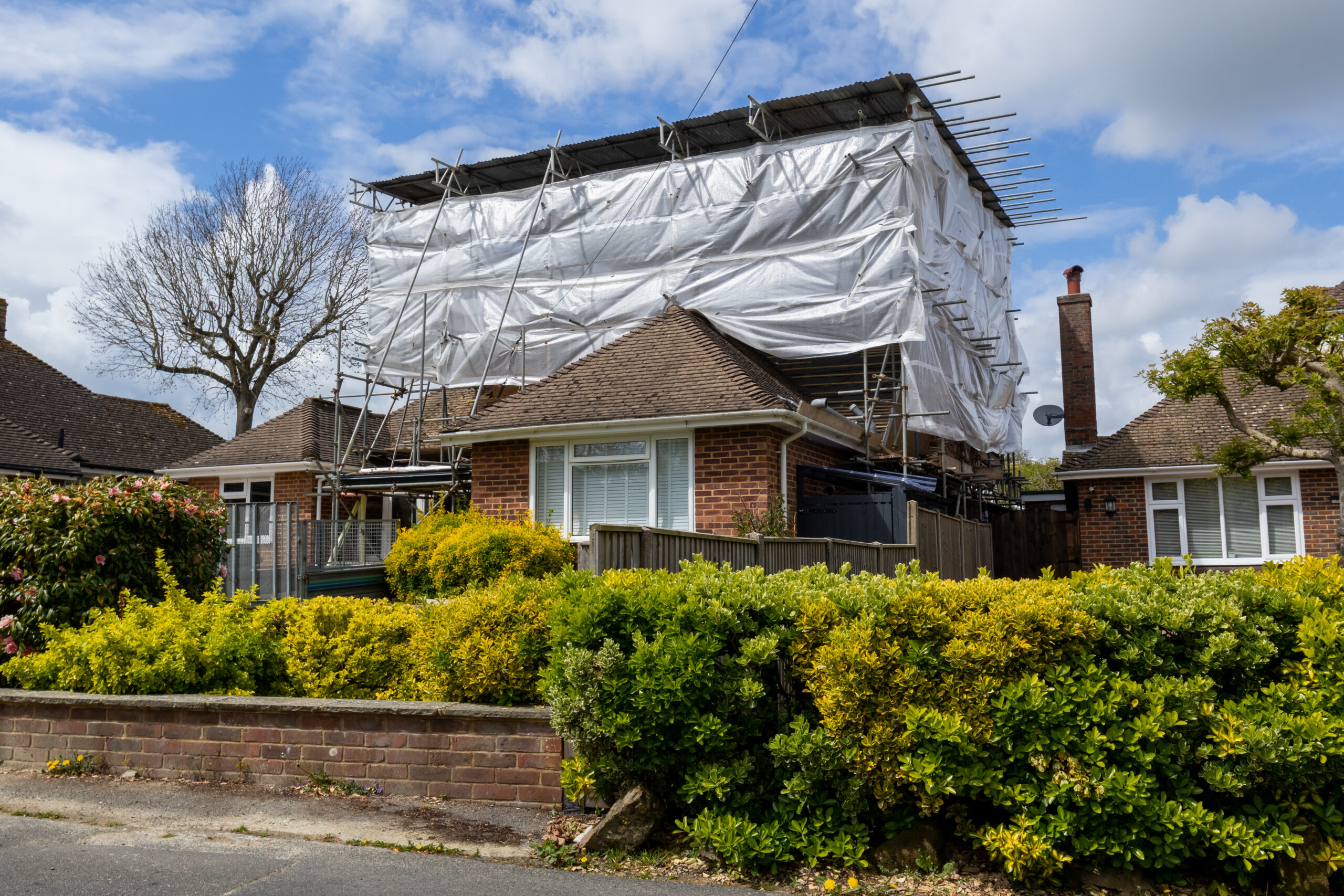 A brick house with a brown roof surrounded by bushes, partially covered in scaffolding and white plastic sheeting for construction or renovation, under a partly cloudy sky.