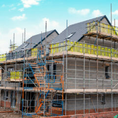 A newly built house surrounded by scaffolding, with unfinished brickwork and roofing under a blue sky, indicating ongoing construction work.