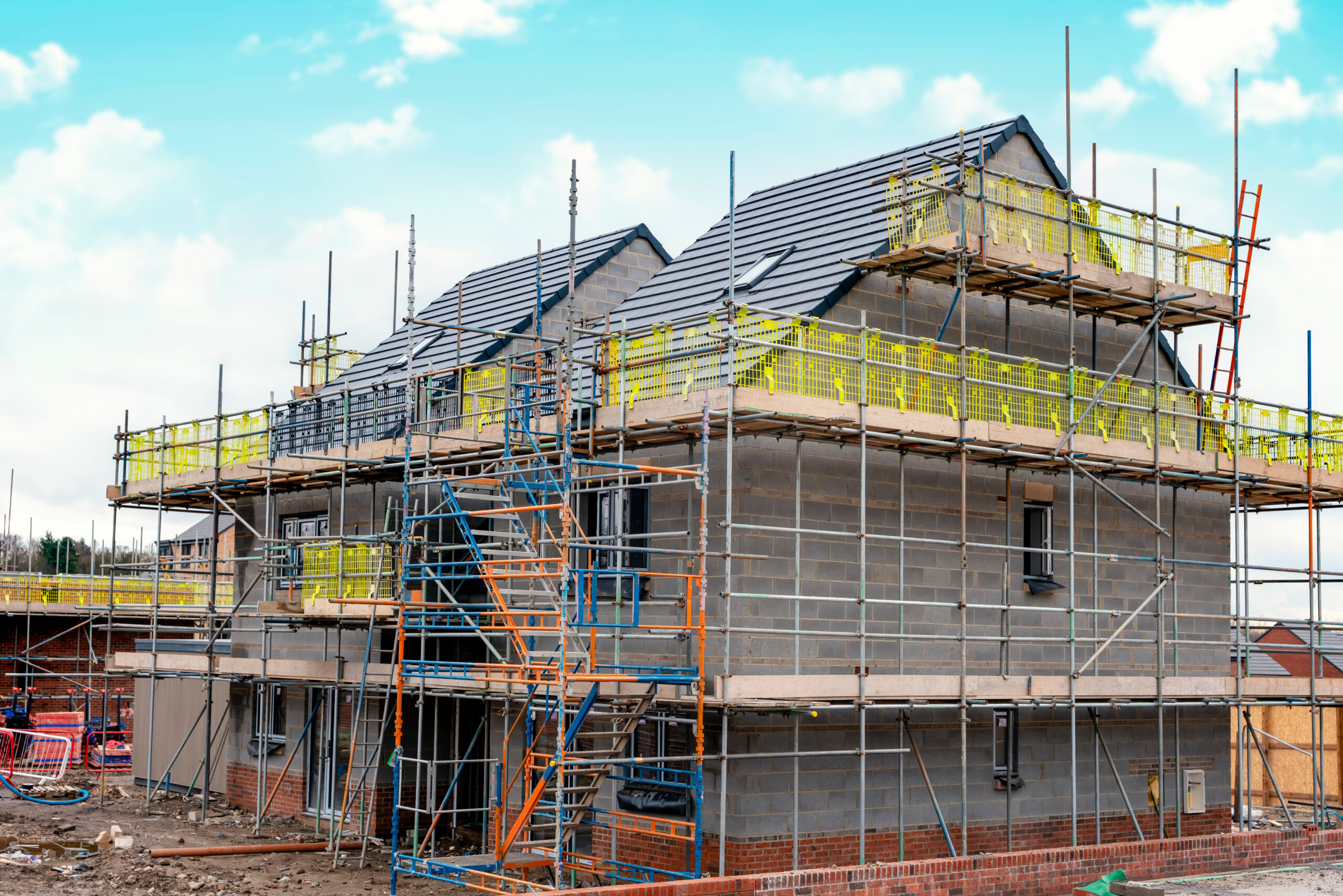 A newly built house surrounded by scaffolding, with unfinished brickwork and roofing under a blue sky, indicating ongoing construction work.