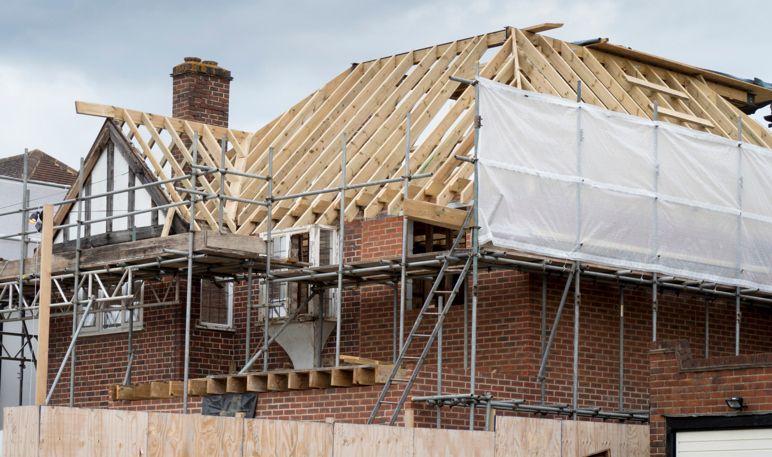 A brick house under renovation with scaffolding and a partially constructed wooden roof frame, surrounded by construction materials and protective sheeting.