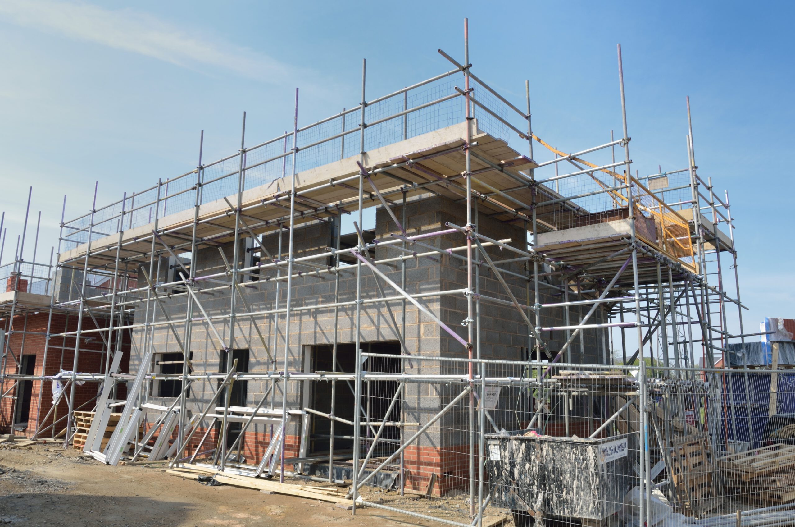 A two-story building under construction with scaffolding around it. The structure has gray concrete blocks and some brickwork, with construction materials and equipment visible on the ground. The sky is clear.