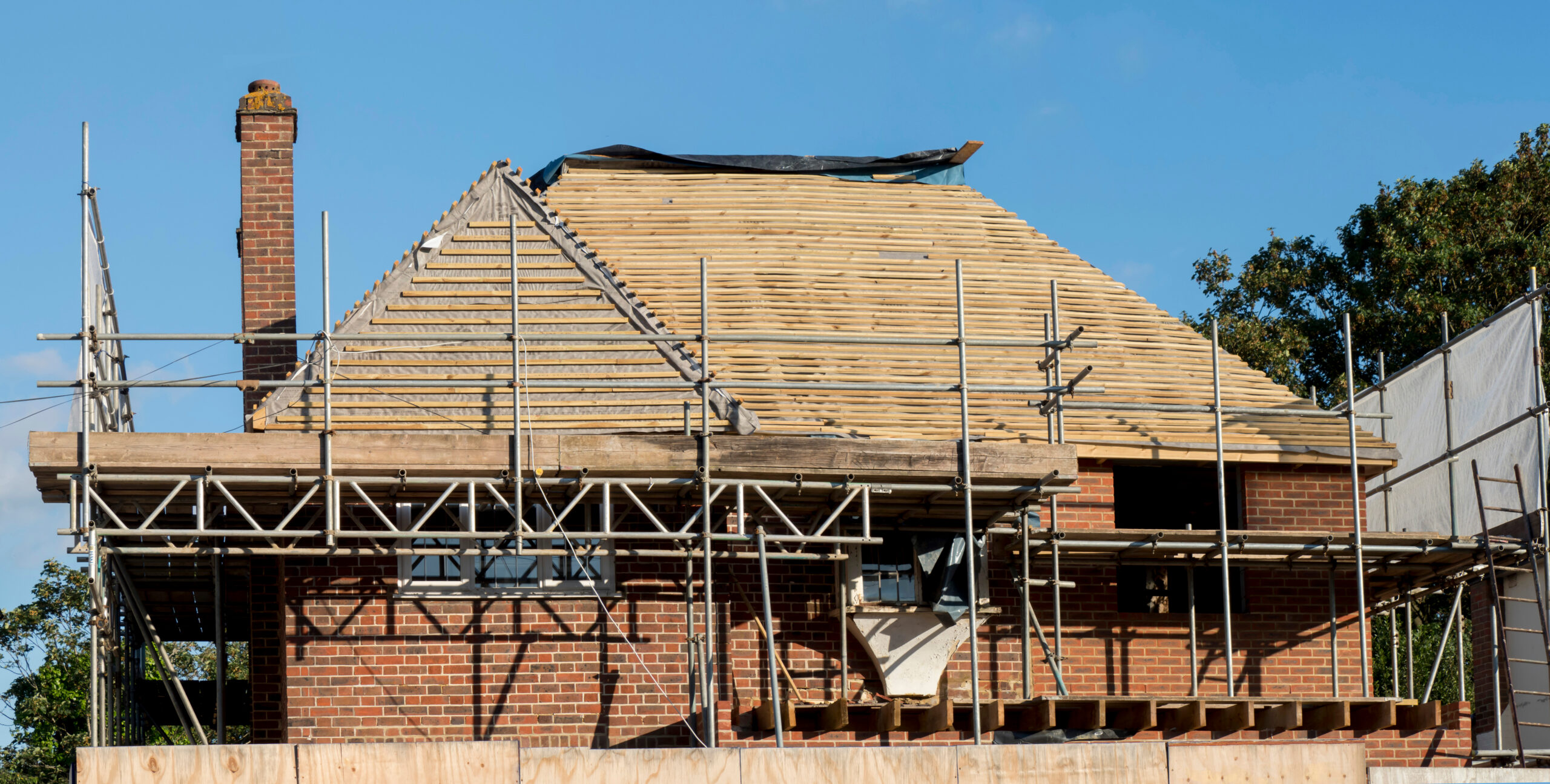 A brick house under construction with scaffolding around it and wooden roof beams exposed, set against a blue sky.