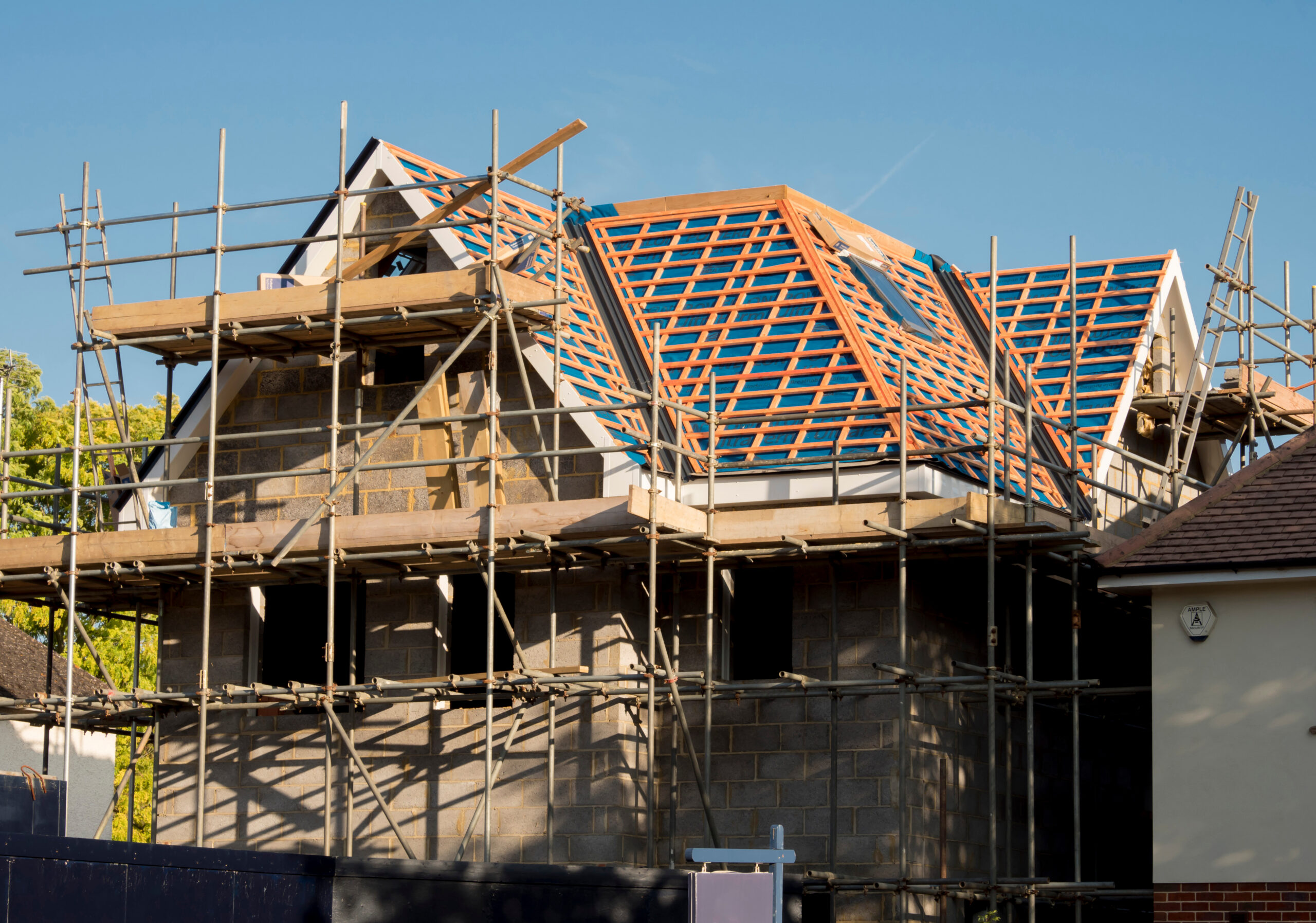 A house under construction with scaffolding around it; the roof frame is exposed, showing wooden beams and blue insulation panels, while the stone walls remain unfinished. The sky is clear and sunny.