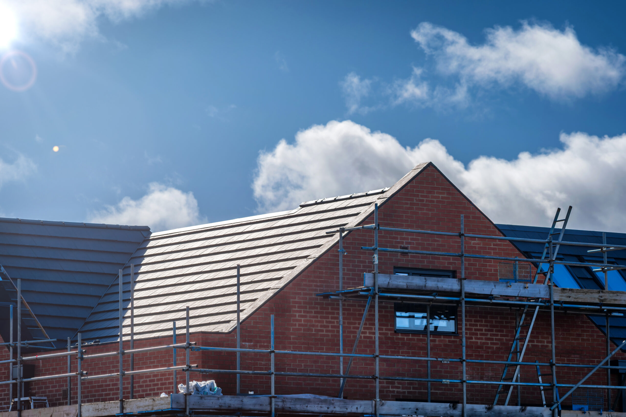 A newly constructed brick house with a tiled roof, surrounded by scaffolding under a blue sky with clouds.