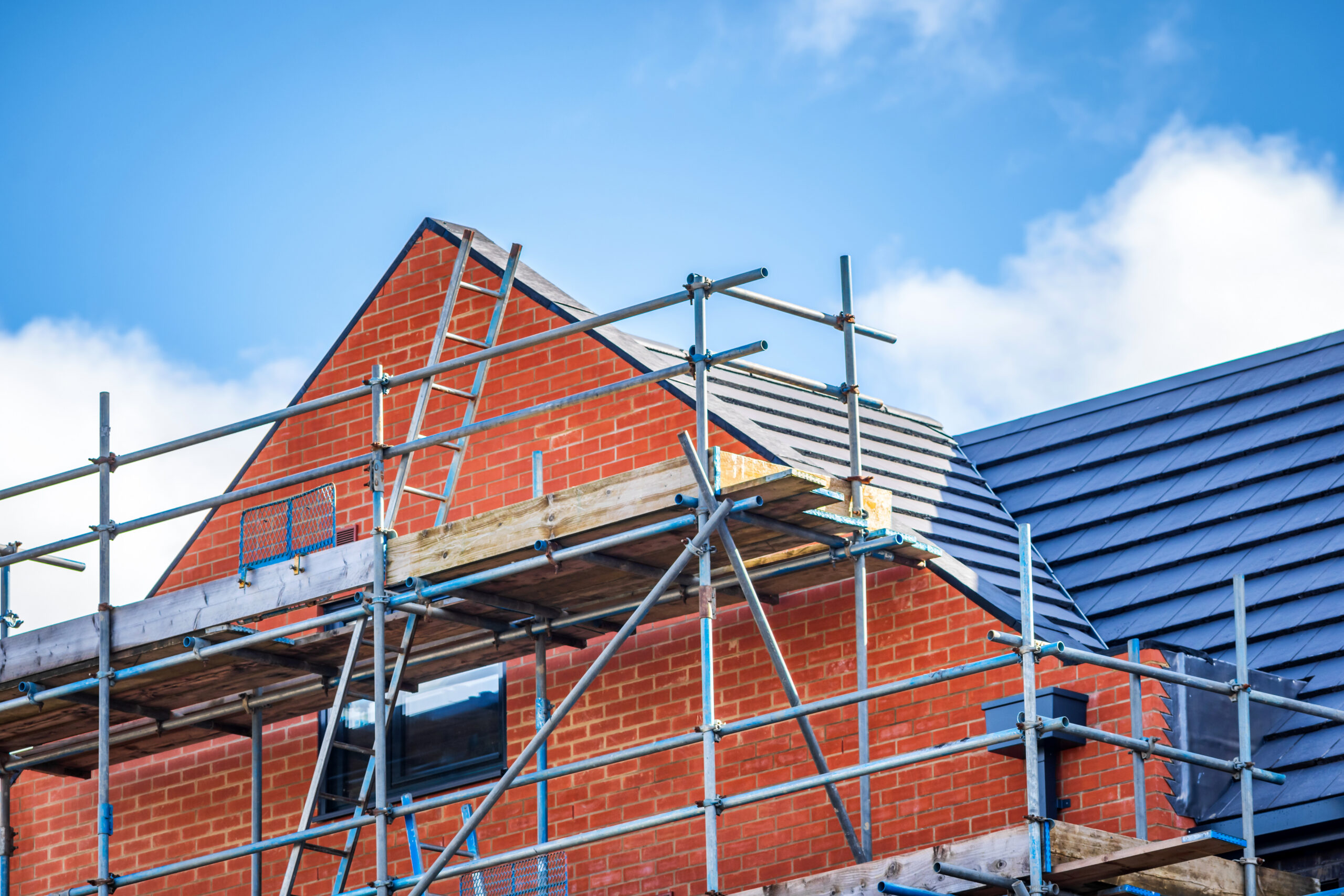 A partially constructed red-brick house with scaffolding around the roof, under a blue sky with scattered clouds.