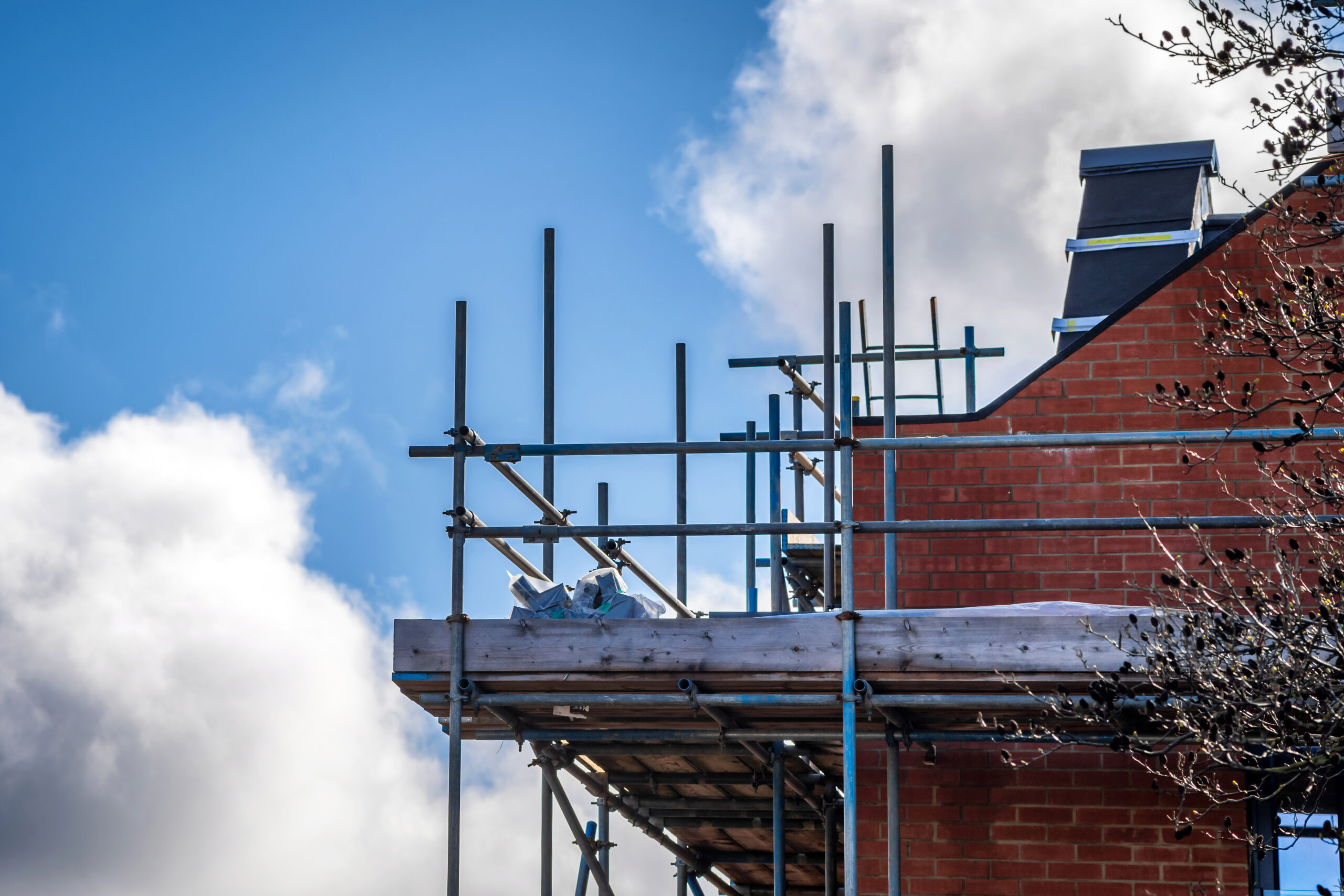 Scaffolding surrounds the upper level of a brick building under construction or renovation, with a partly cloudy sky in the background and some tree branches visible on the right.