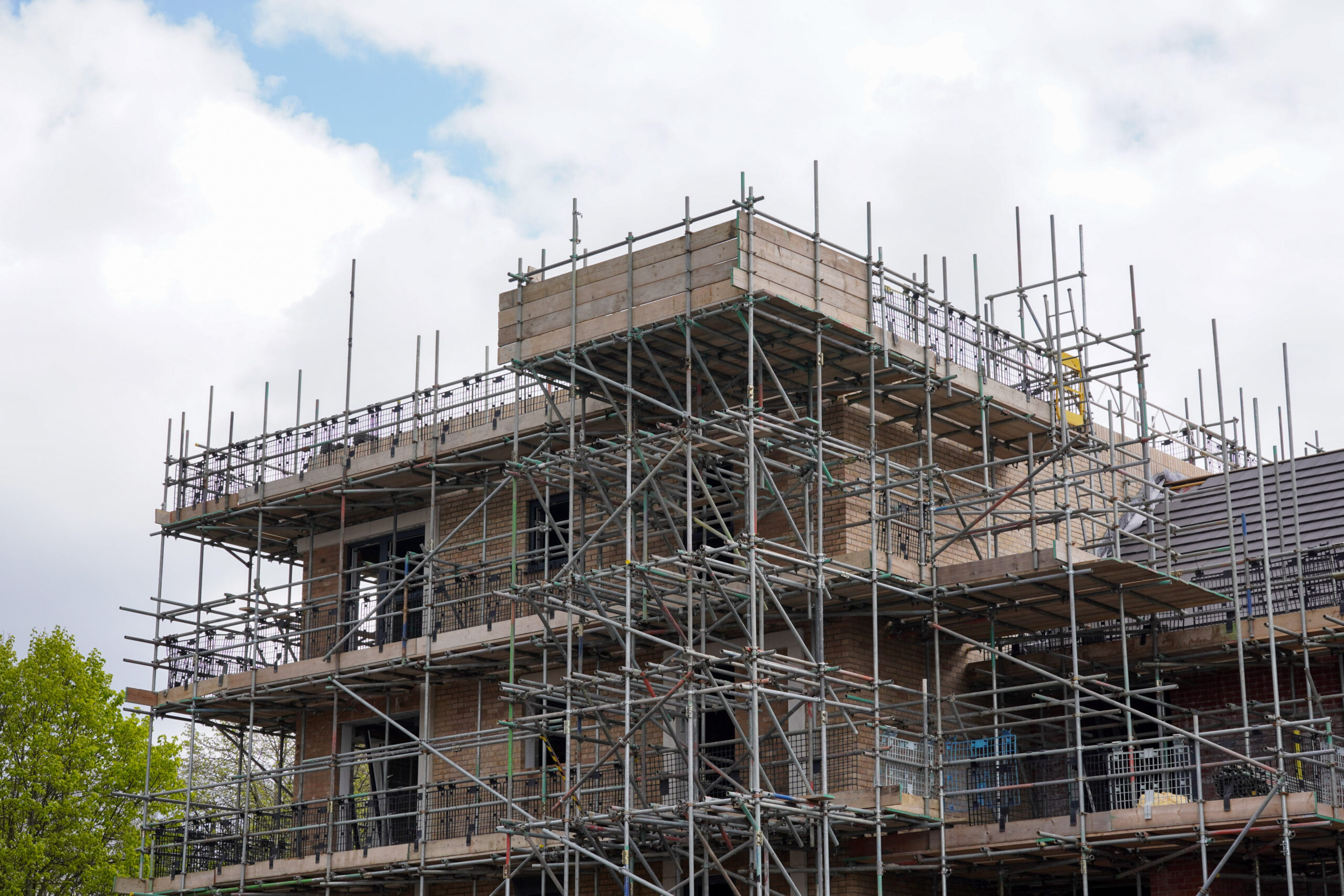 A multi-story building under construction, surrounded by extensive metal scaffolding, with some completed walls and open window spaces visible under a partly cloudy sky.