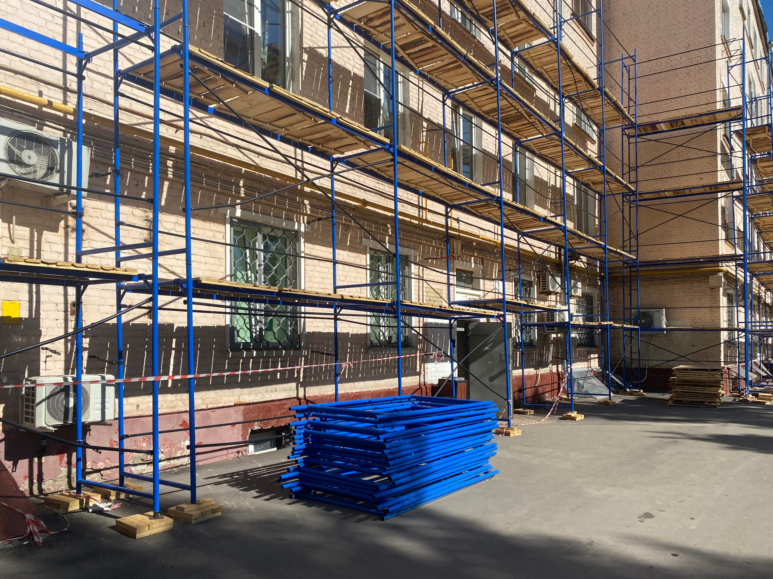 Blue scaffolding is set up along the side of a brick building, with stacked blue metal frames on the ground nearby. The area is sunny, and the building has bars on the windows and air conditioning units attached.