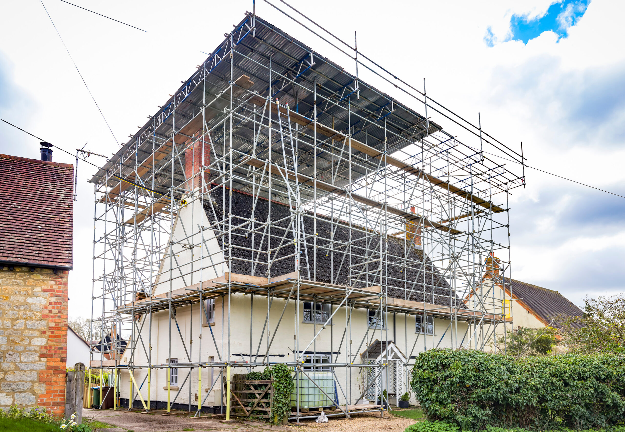 A white house with a steep roof is surrounded by extensive scaffolding, indicating construction or renovation work. The sky is partly cloudy, and there are other houses and greenery nearby.