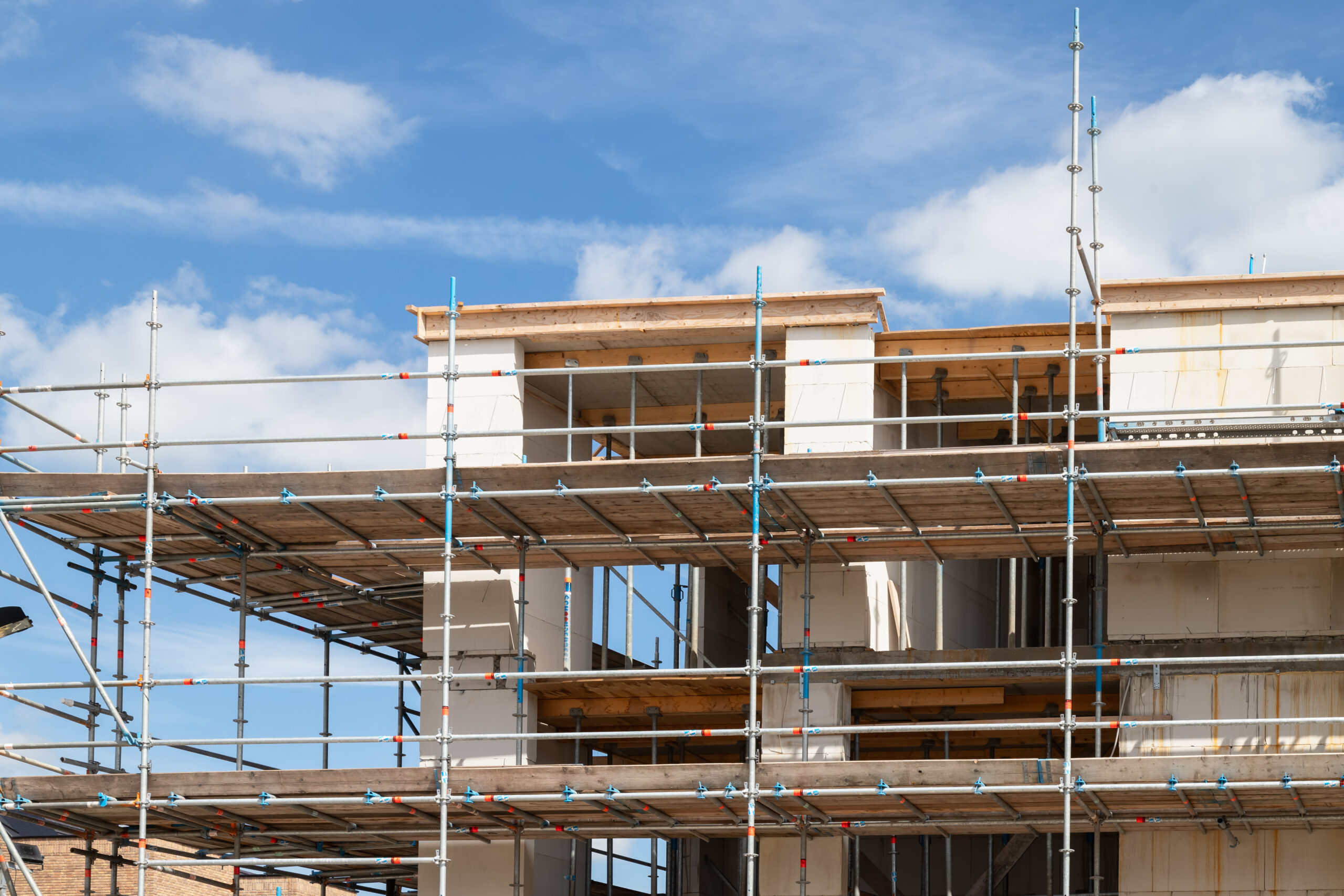 A partially constructed building with exposed concrete walls and wooden beams, surrounded by metal scaffolding under a blue sky with scattered clouds.