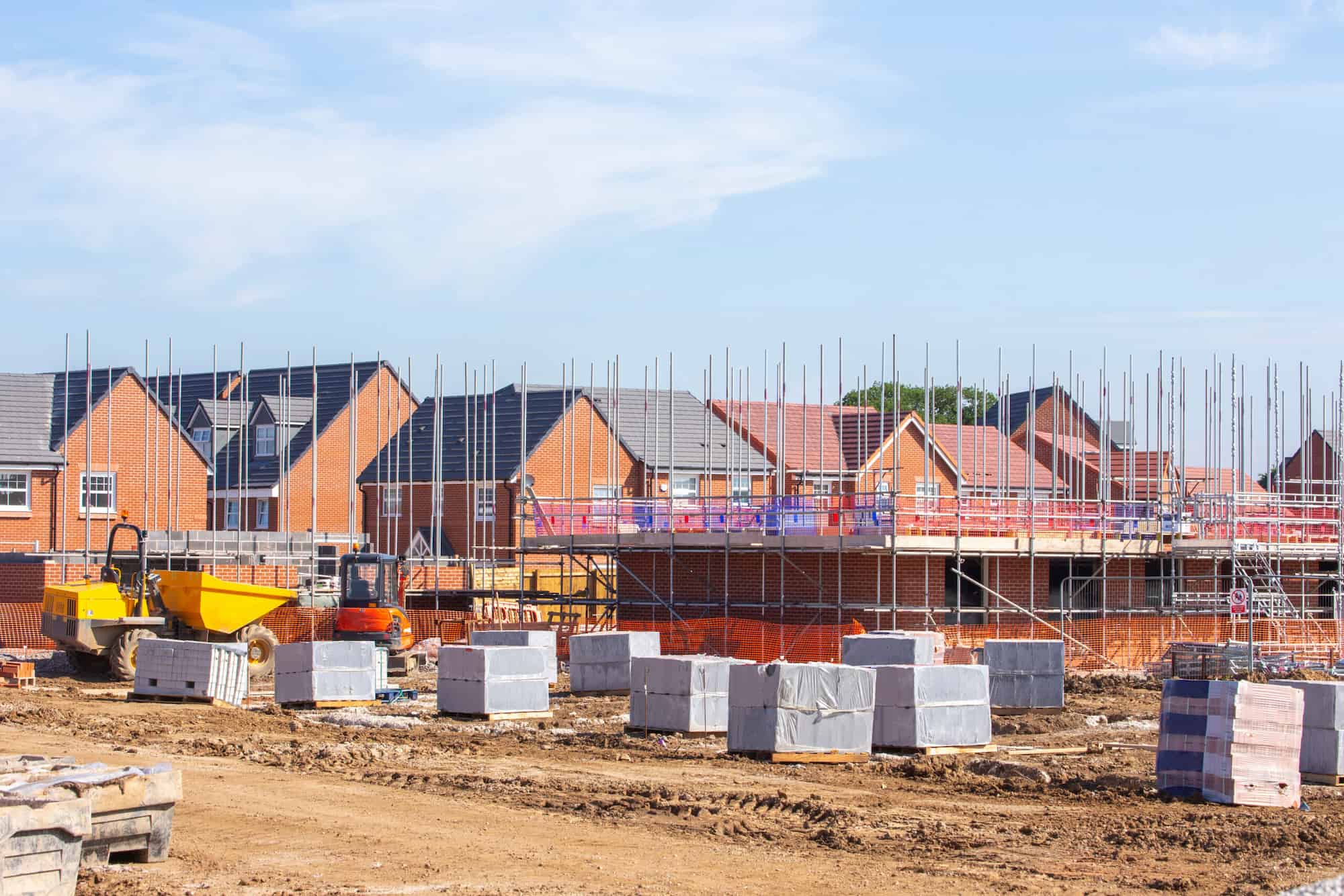 A housing construction site with partially built brick houses, scaffolding, construction vehicles, and stacks of building materials under a blue sky.