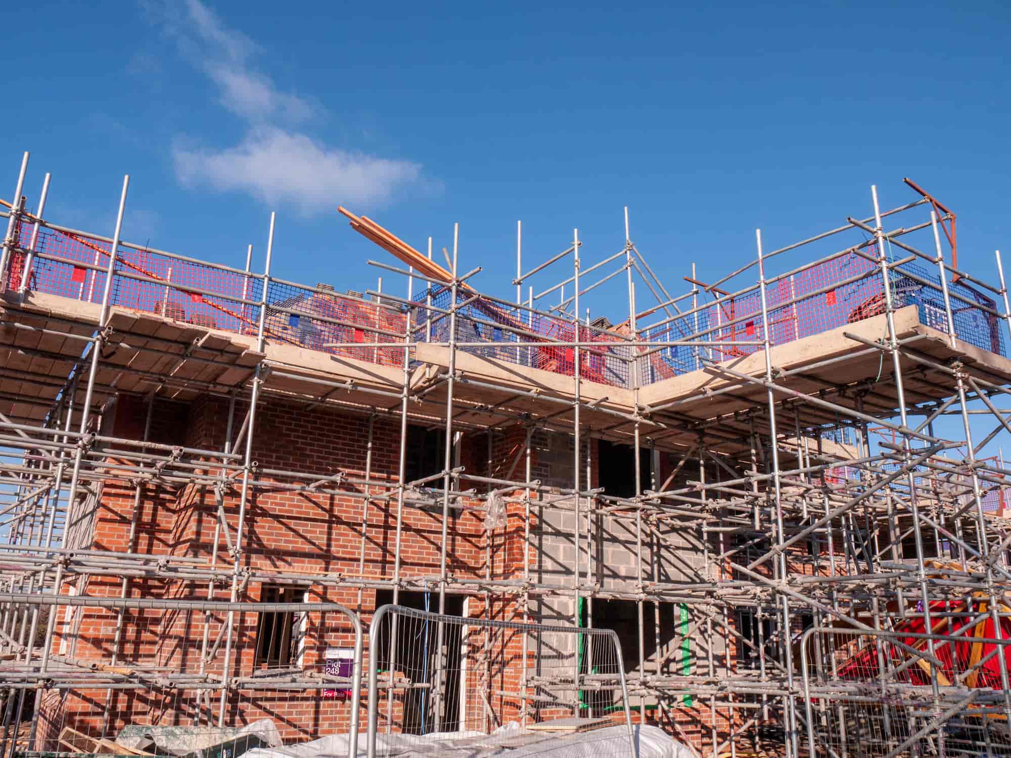 A brick building under construction, surrounded by metal scaffolding and netting, against a clear blue sky. Construction materials and tools are visible on the upper level.