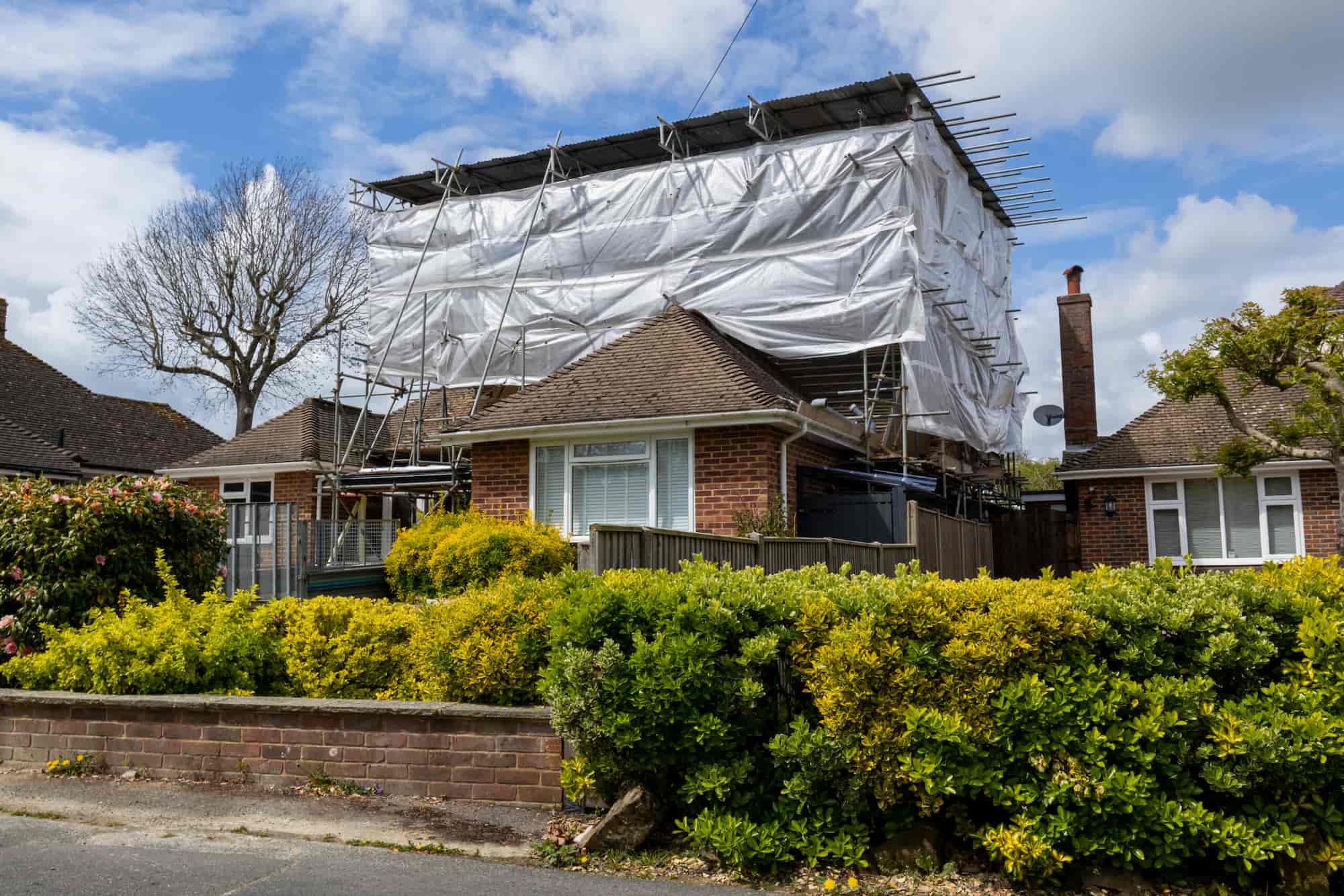 A brick house with scaffolding and white plastic sheeting covering its roof, surrounded by greenery and bushes, under a partly cloudy sky.