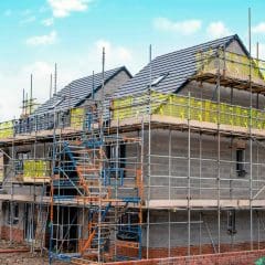 A partially constructed house with scaffolding around it, yellow safety netting, and construction materials visible. The sky is blue with some clouds in the background.