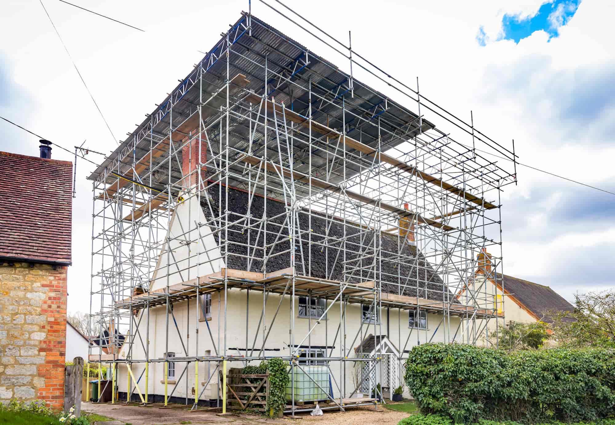 A house surrounded by extensive scaffolding, covering the roof and sides, suggesting construction or renovation work. The sky is partly cloudy, and there is greenery in the foreground.